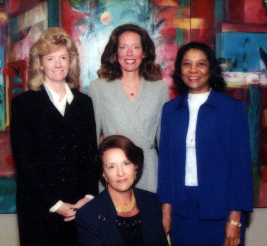 Image Description. Photograph of three women standing, two who are Caucasian and one woman of color. A Caucasian woman sits posed in front of the other three.