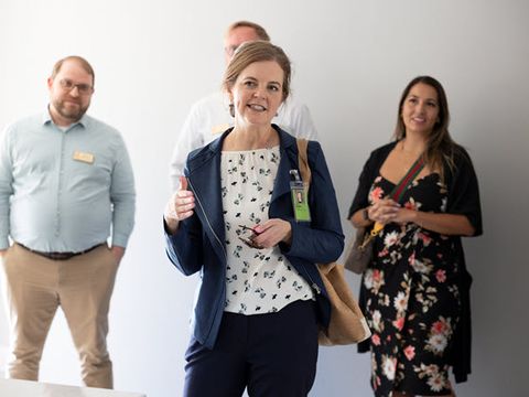 From left to right, we have a white man wearing khaki pants and a blue shirt, a white woman in a black pantsuit and white blouse, who is talking, and a white woman with long brown hair wearing a floral print dress.