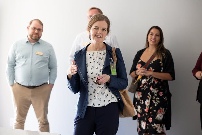 From left to right, we have a white man wearing khaki pants and a blue shirt, a white woman in a black pantsuit and white blouse, who is talking, and a white woman with long brown hair wearing a floral print dress.