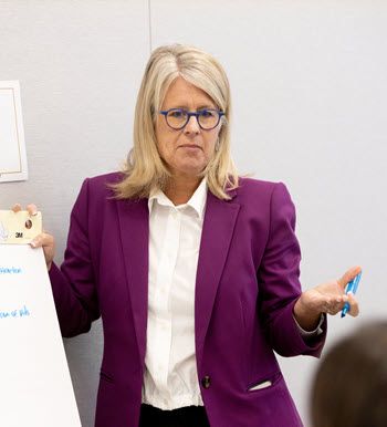 A White woman with blonde, shoulder-length hair and glasses stands at a flip chart. She's wearing a white shirt and magenta jacket, and holding a blue marker.