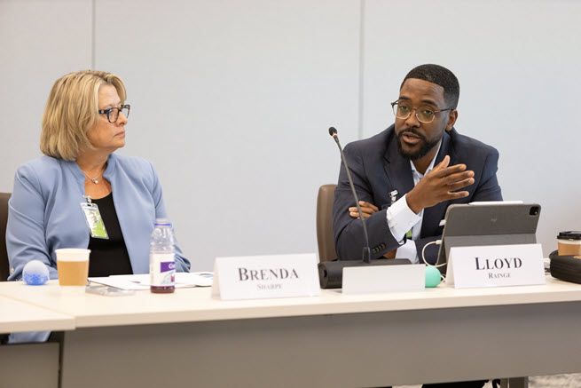 To the left is a White women with blonde hair and classes wearing a light blue jacket and navy shirt. She is listening to a Black man with glasses wearing a gray suit and blue shirt who is seated to her left.