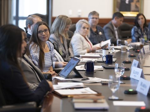 A young woman turns towards the camera, in conversation with her neighbor. She is along a row of about eight people along the side of a big wooden conference table.