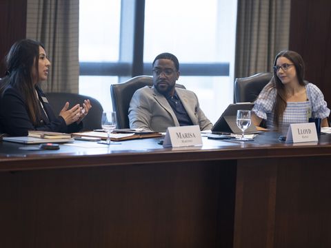 A Latinx woman with long dark hair is speaking at a large conference table. A Black man in a gray suit and blue shirt and a White woman in a gingham dress are listening attentively.