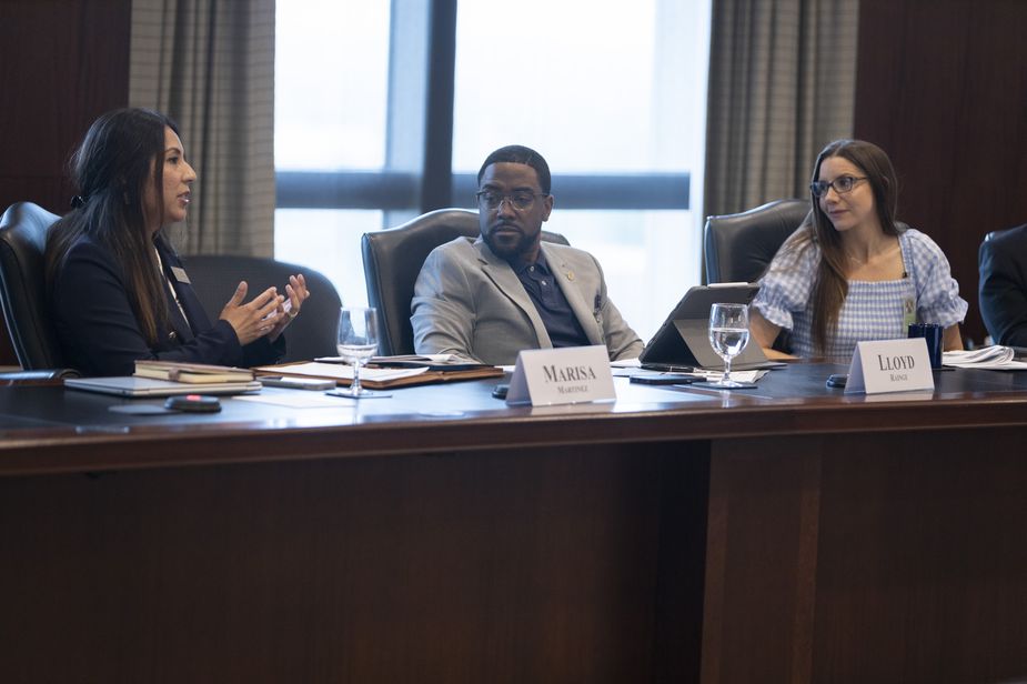 A Latinx woman with long dark hair is speaking at a large conference table. A Black man in a gray suit and blue shirt and a White woman in a gingham dress are listening attentively.