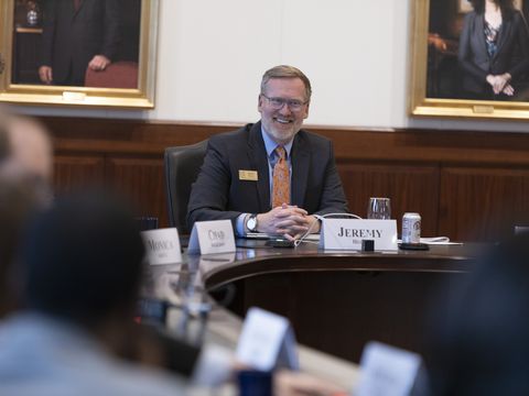 At the head of the conference table, a smiling, bearded White man in a dark suit and red tie, faces the camera.