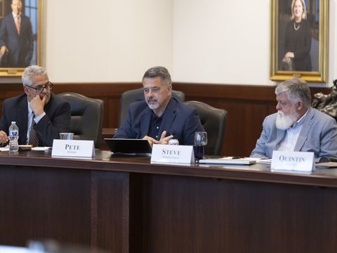 The picture includes three men seated at a large wooden conference table. The man in the middle is Native American. He has dark brown hair with some gray and is wearing a blue suit, and he's talking. The man to the left is Latinx, with gray hair and glasses, wearing a gray suit. The man to the right is White, with a long white beard and white hair, and a light blue suit.