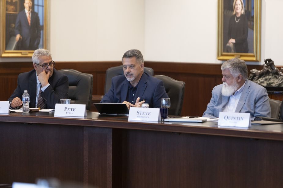The picture includes three men seated at a large wooden conference table. The man in the middle is Native American. He has dark brown hair with some gray and is wearing a blue suit, and he's talking. The man to the left is Latinx, with gray hair and glasses, wearing a gray suit. The man to the right is White, with a long white beard and white hair, and a light blue suit.