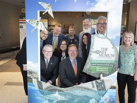 The Money Museum has a big frame where people can post behind the opening for a photo. The group posing today includes ten people, six men and four women. They're smiling, kind of goofy, and appear to be enjoying themselves.