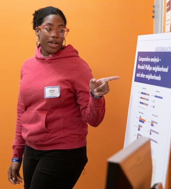 A young Black woman in pink glasses and a pink hoodie gestures to a large board that has a map and data on it. She's the facilitator, and she's leading a group discussion.