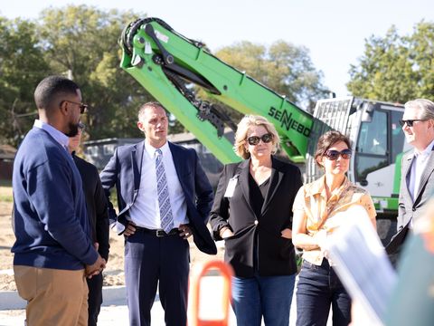 Five people occupy the front of the photo, while a gigantic green metal excavator looms over them. The people include a black man in a blue jacket, a white man in a suit and tie, a blonde woman in a black knit jacket, a brown-haired woman in a yellow shirt, and a white man in sunglasses and a suit.