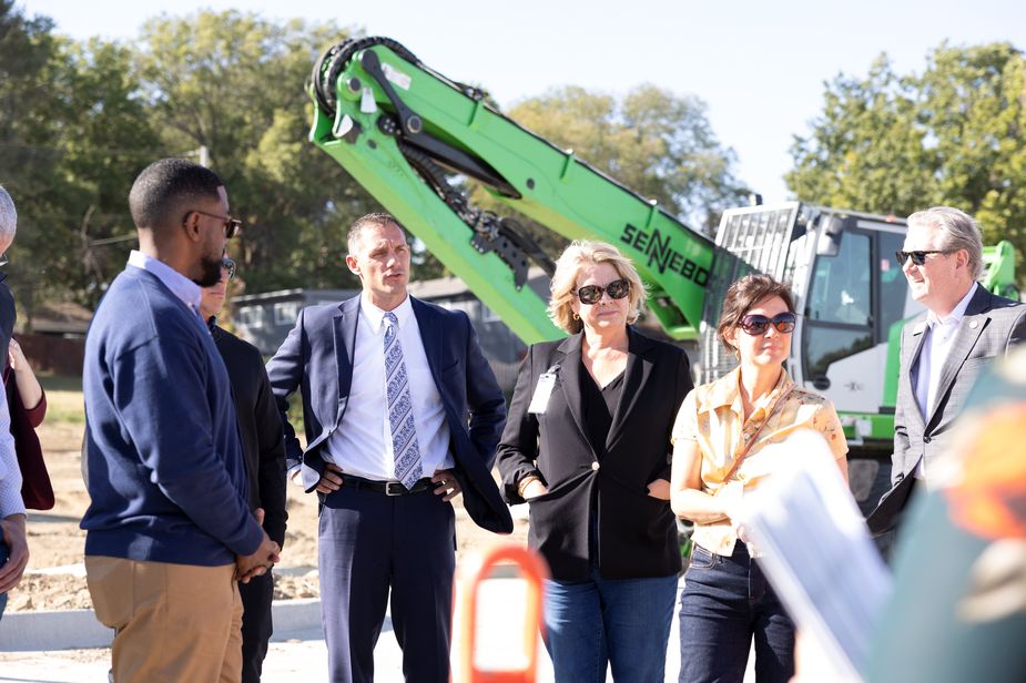 Five people occupy the front of the photo, while a gigantic green metal excavator looms over them. The people include a black man in a blue jacket, a white man in a suit and tie, a blonde woman in a black knit jacket, a brown-haired woman in a yellow shirt, and a white man in sunglasses and a suit.