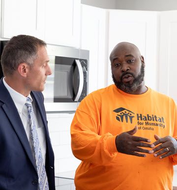 A white man in a gray business suit, white shirt and light gray tie is leaning toward a black man wearing a bright orange t-shirt that says Habit for Humanity, who is talking.