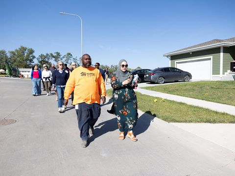 A black man in a bright orange t-shirt and a white woman in a hijab and floral pantsuit are at the front of a group of people walking through a subdivision.