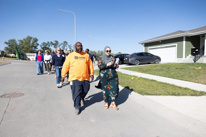 A black man in a bright orange t-shirt and a white woman in a hijab and floral pantsuit are at the front of a group of people walking through a subdivision.