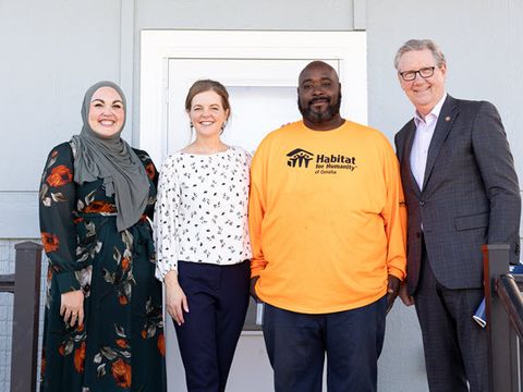 From left to right, we see a woman wearing a hijab and flowered dress, a white woman with brown hair wearing black pants and a white blouse, a black man in an orange t-shirt and a white man wearing glasses and a gray suit.