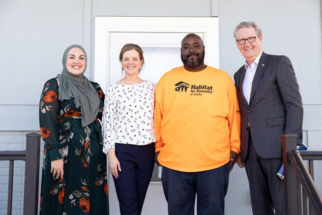 From left to right, we see a woman wearing a hijab and flowered dress, a white woman with brown hair wearing black pants and a white blouse, a black man in an orange t-shirt and a white man wearing glasses and a gray suit.