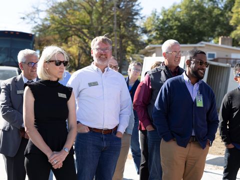 A group of people are outside, facing a construction site. From left to right, we have an Hispanic man in a gray suit, a blonde woman in a black shirt and pants, a white man wearing a blue shirt and jeans, a white man in a vest and long-sleeved t-shirt, a black man in a navy cardigan and khaki pants, and an Hispanic man in sunglasses, sweatshirt and jeans.