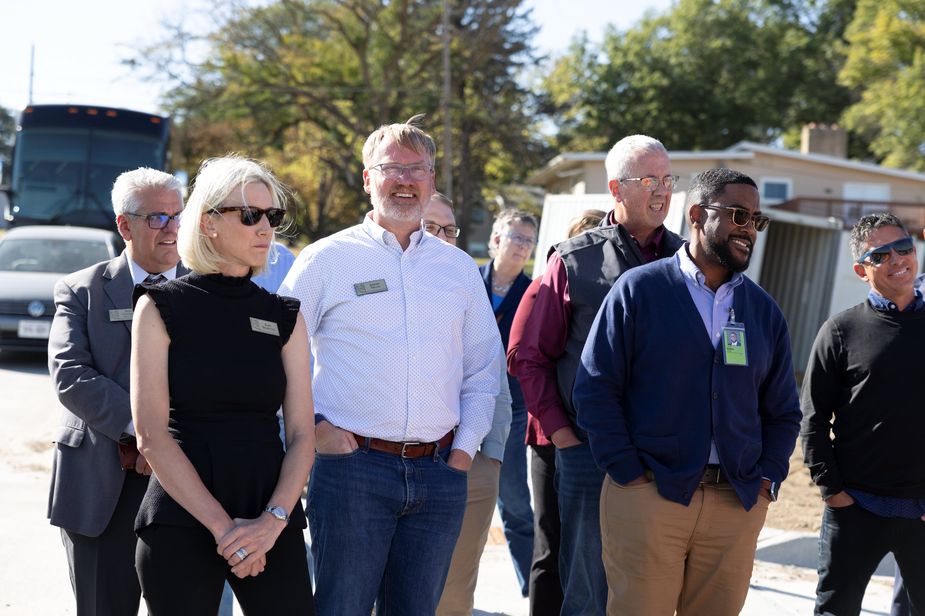 A group of people are outside, facing a construction site. From left to right, we have an Hispanic man in a gray suit, a blonde woman in a black shirt and pants, a white man wearing a blue shirt and jeans, a white man in a vest and long-sleeved t-shirt, a black man in a navy cardigan and khaki pants, and an Hispanic man in sunglasses, sweatshirt and jeans.