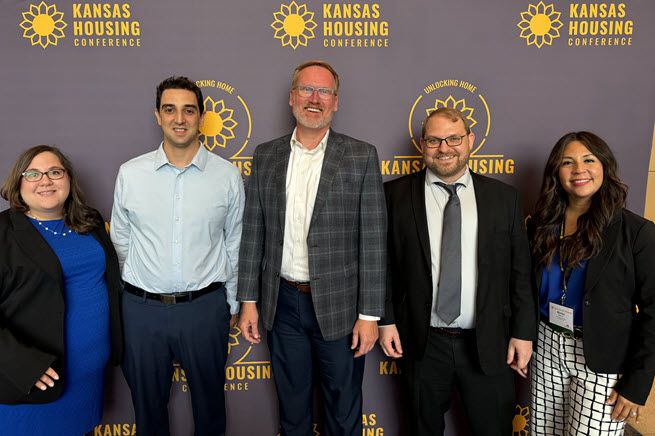 The photo is of five people from the Kansas City Fed. The woman on the far left is in her 30s, wearing glasses and a midnight blue dress and black blazer. Next to her is a dark-haired man in his 30s wearing a light blue shirt and black pants. He's next to a blonde man with a beard, in his late 40s, wearing a white shirt and gray suit. Next is another bearded man, this time with brown hair, and wearing a dark suit. At the far right is a woman with long, dark hair, wearing a midnight blue shirt and black blazer.