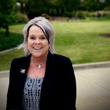 Smiling woman standing outside in a park. She's wearing a blue dress and black sweater and has steel-gray hair.