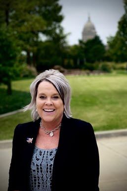 A smiling white woman in a blue dress and black blazer stands outside in front of what appears to be the state capitol of Oklahoma.