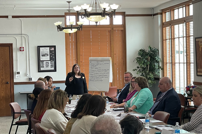 A woman in a black dress is smiling as she stands by a flip chart at the head of a long table. Each seat is filled, and she is encouraging everyone to participate.