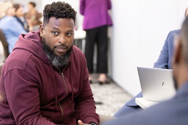 The photo shows a Black man with a beard, wearing a magenta hoodie.