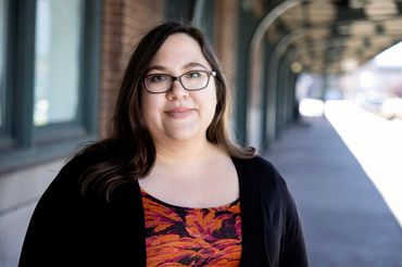 A woman in her 30s with long brown hair and glasses is in the foreground, standing in front of what appears to be an historic railroad station. She's wearing a black sweater over a dark floral dress.