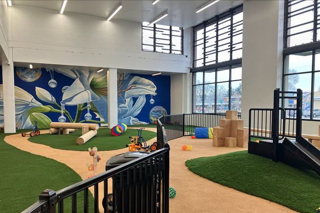 The childcare center has a light brown path through bright green turf, and murals in blue tones on the walls. Floor-to-ceiling windows create a bright space.