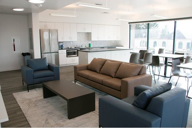 A spacious community room has teal and brown sofas, a wooden coffee table, and a small kitchen with new appliances. French doors bring in natural light.