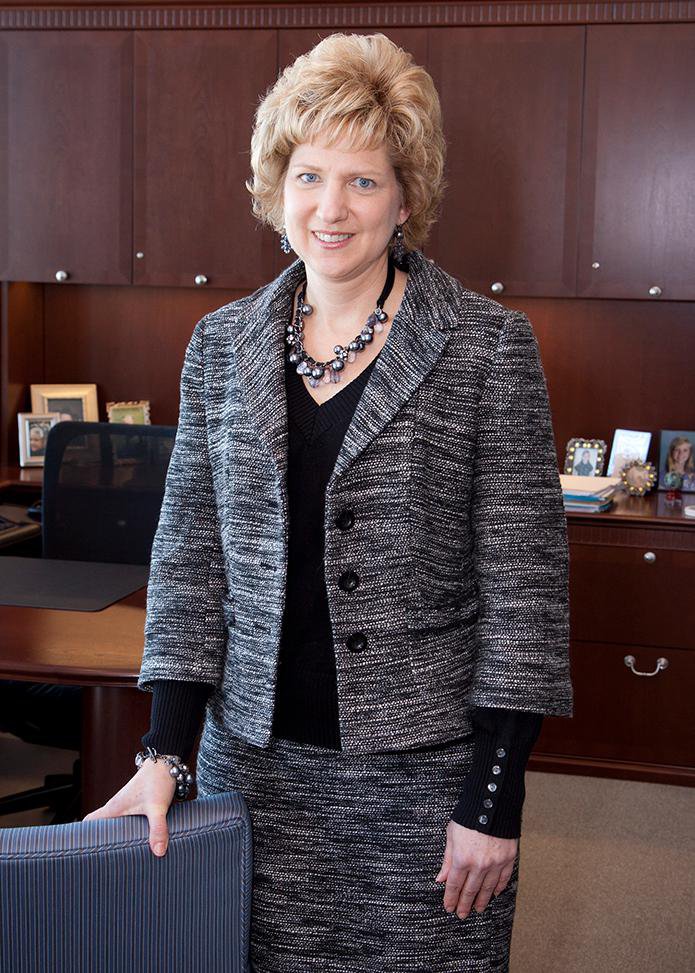 Image Description. Photograph of a Caucasian woman with short blond hair standing in an office, smiling.